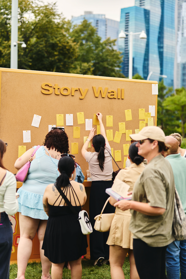 People interacting with a community story wall covered in notes in an outdoor setting.