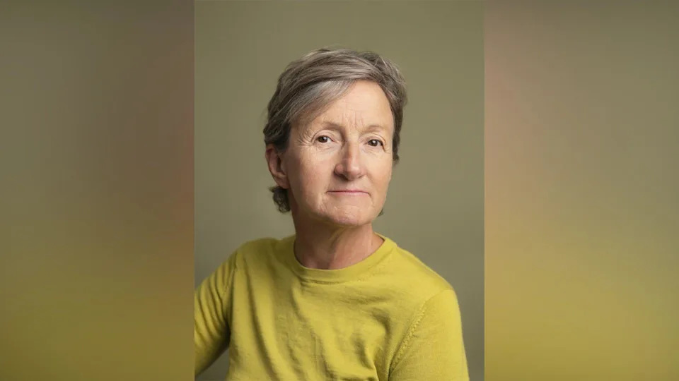 Sarah Hendrickx. She has short grey hair and brown eyes. She is wearing an acid green jumper and looking at the camera with a slight smile.