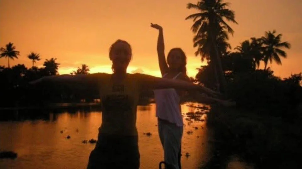 Lowri and a friend dancing in the sunset in India. They stand in front of a lake with palm trees on the banks.