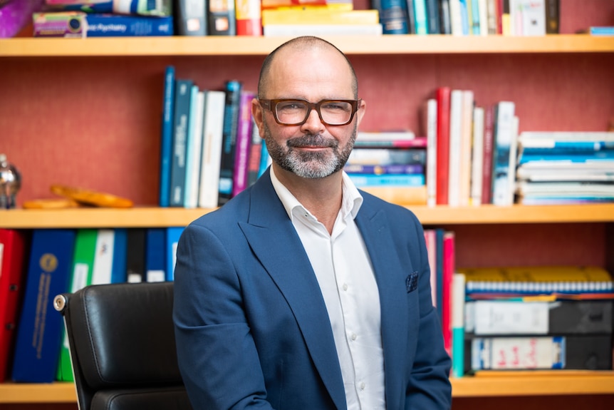 A man wearing a suit and glasses looks at the camera, he is standing in front of a shelf of colourful books.