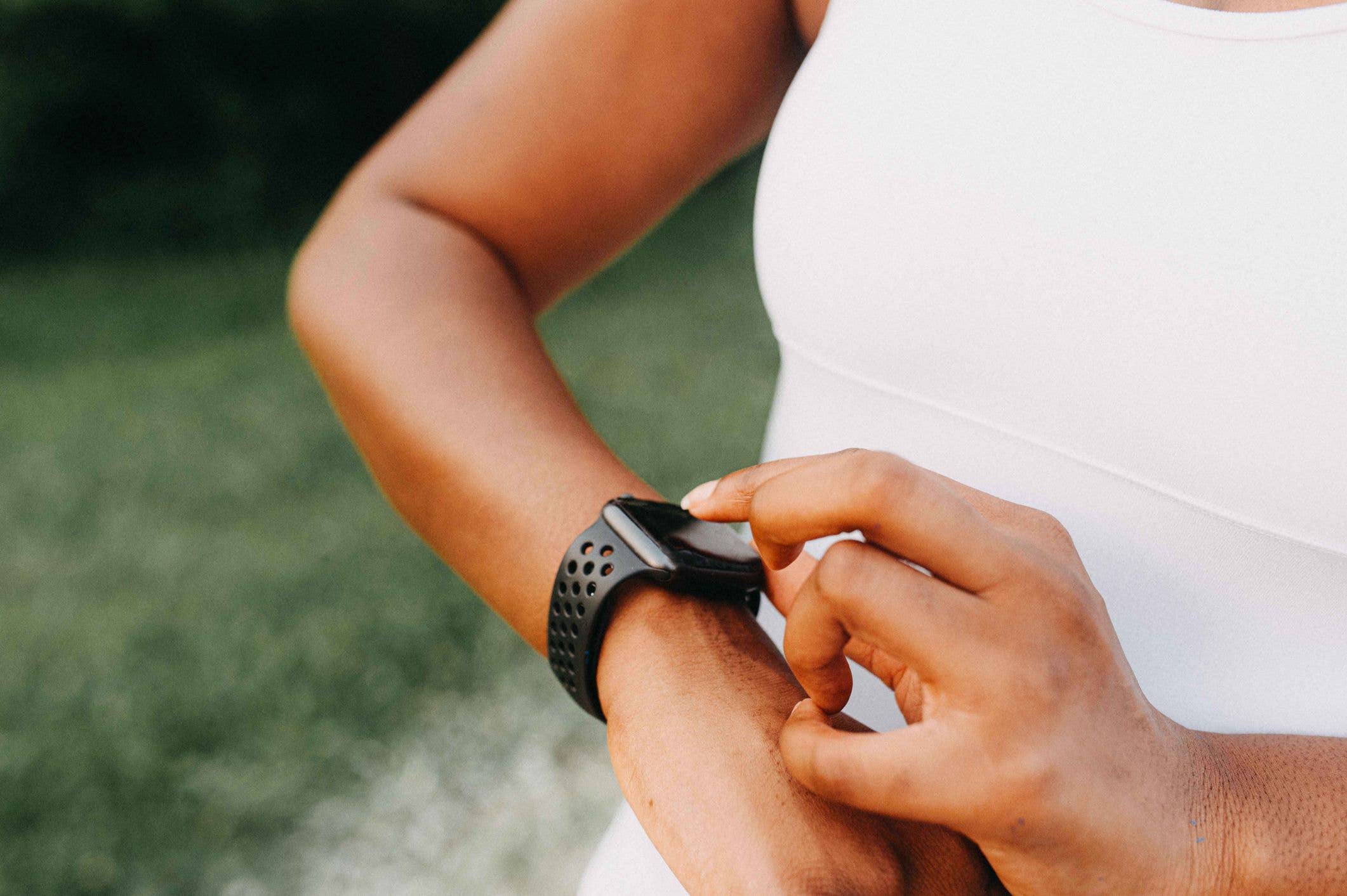 A woman's hand is interacting with a smart watch on her wrist. The outdoor setting has a blurred green background, emphasizing the focus on the wearable technology.