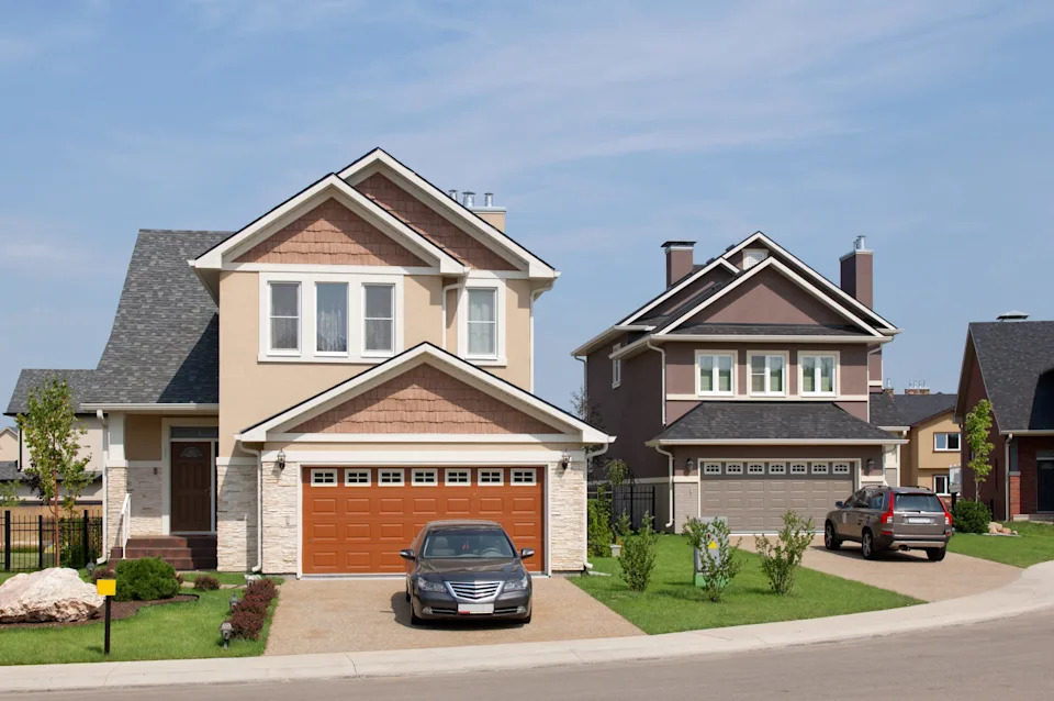 Two suburban houses with cars parked in their driveways, featuring manicured lawns and a clear sky in the background