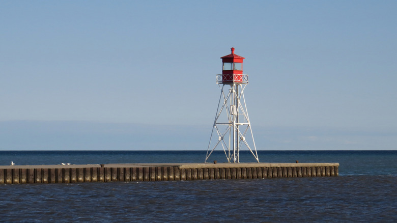 An open framework style lighthouse sits at the end of a concrete pier; a small red box-like structure sits on top which houses the lighthouse light
