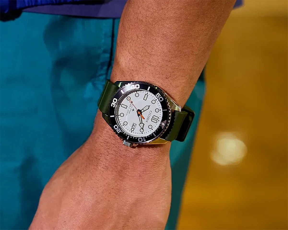 Wristwatch with white dial, black bezel, and green strap worn on a wrist.