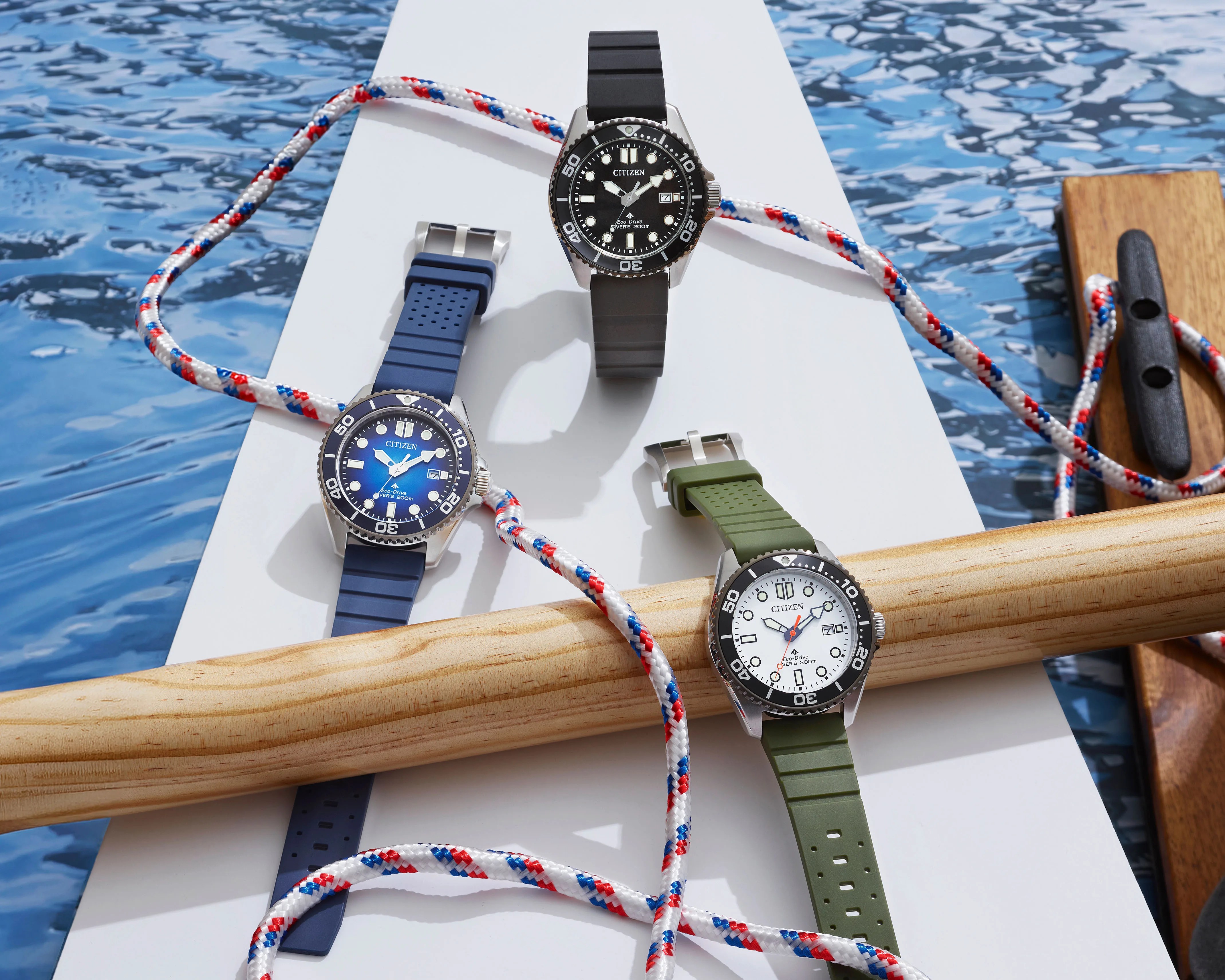Three Citizen Eco-Drive diver watches with black, blue, and white dials and rubber straps in black, blue, and green on a white surface with nautical rope and wooden oars.