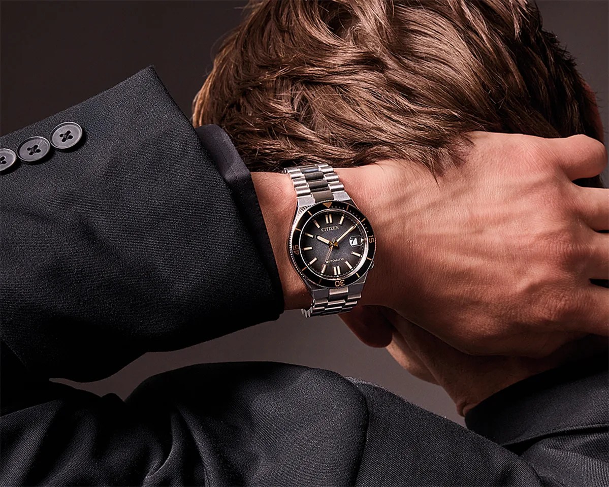 Silver metal Citizen wristwatch with black dial and date window worn on a wrist with a black suit jacket.