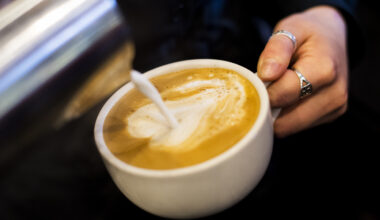 A hand pours milk into a cup of coffee, creating a swirl pattern on the surface.