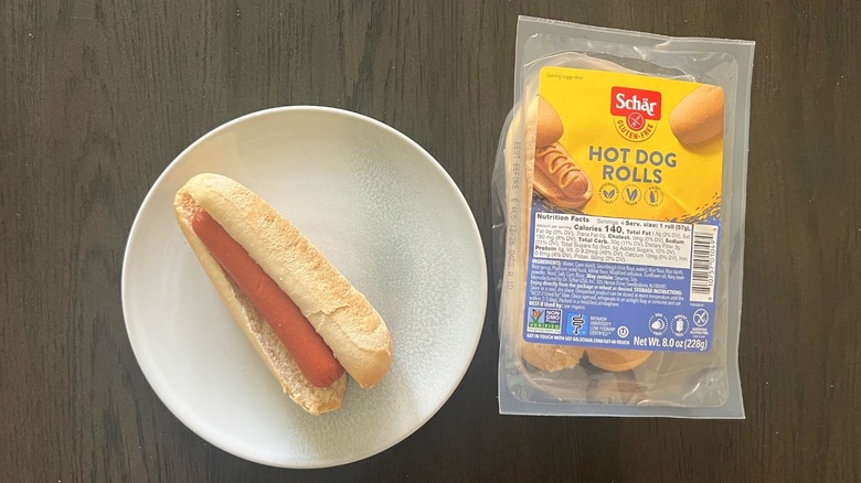 A package of Schär hot dog rolls next to a hot dog on a plate