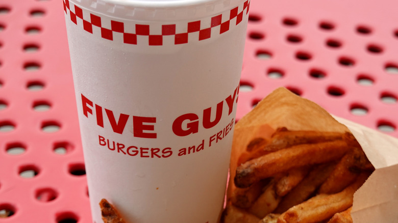 A Styrofoam cup with the 'Five Guys Burgers and Fries' logo on it sits next to a bag of Five Guys fries on a red table