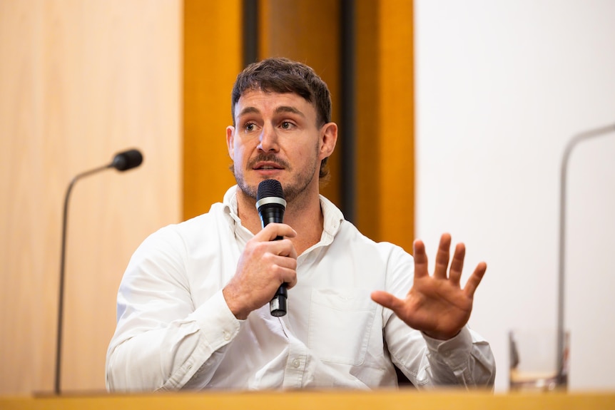 Luke Bateman wears a white-collared shirt seated, holding a microphone, his hand in the air as he speaks.