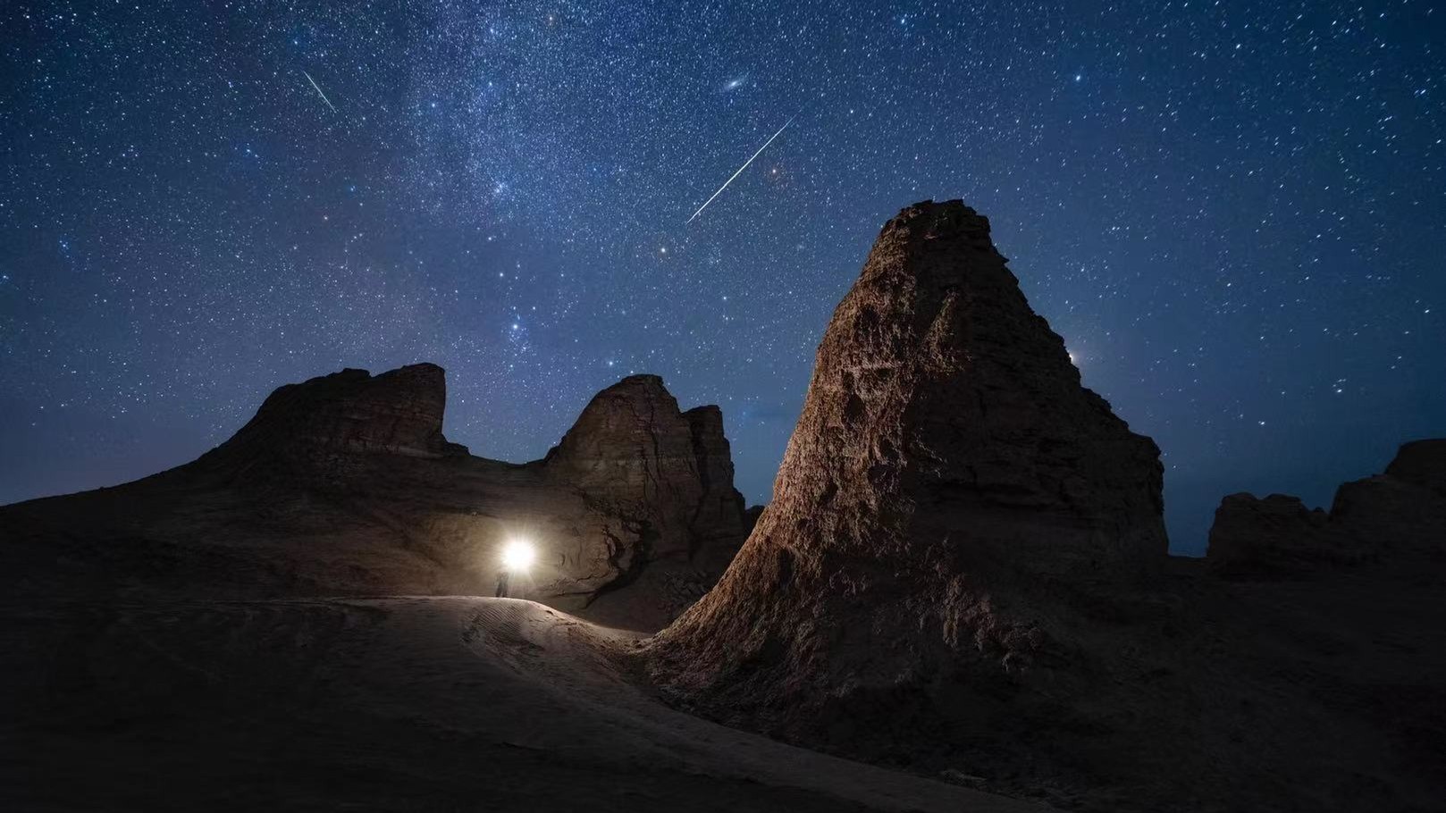 a long meteor streaks through the sky a person below shines a flashlight.