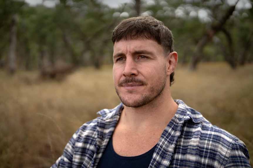 Luke Bateman looks forward, neutral facial expression. He wears a flannelette shirt and stands in dry grassland.
