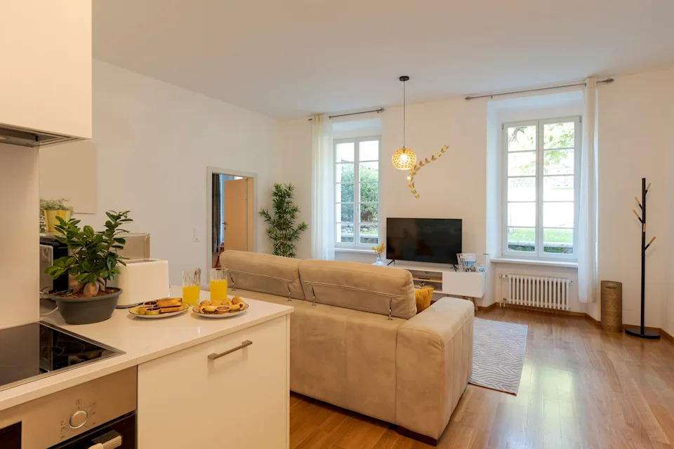 Cozy modern living room with a beige sofa, TV, and two large windows. Open kitchen area with a countertop and orange juice glasses