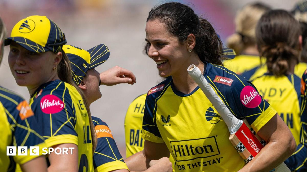 Maia Bouchier in yellow shirt, helmet and pads, and navy trousers, drives with Lancashire wicket and bowler, in all red, also in shot