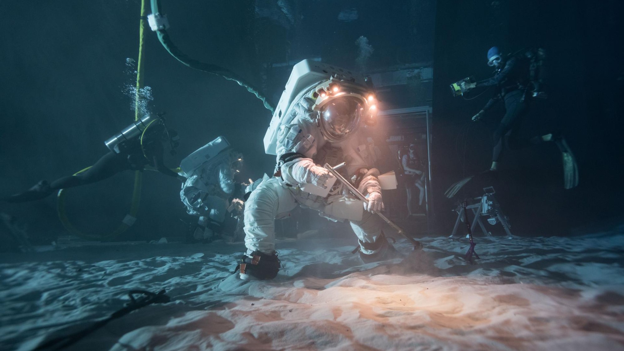a person in a bulky white spacesuit attached to an umbilical digs in sand with a shovel underwater