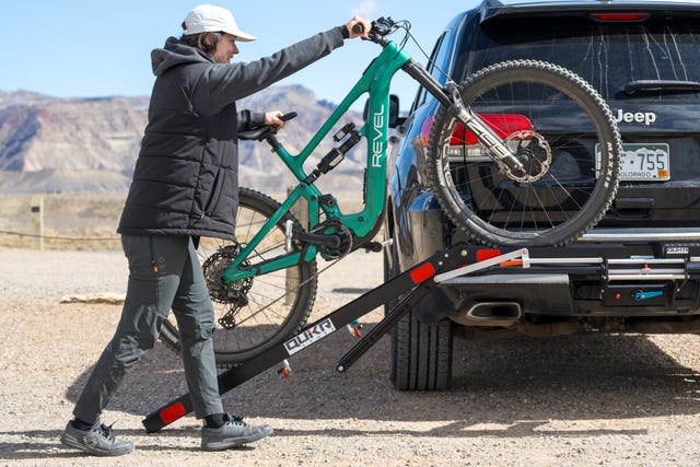 Person loading a green mountain bike onto a bike rack attached to a black SUV in a desert landscape. Person loading a green mountain bike onto a bike rack attached to a black SUV in a desert landscape.