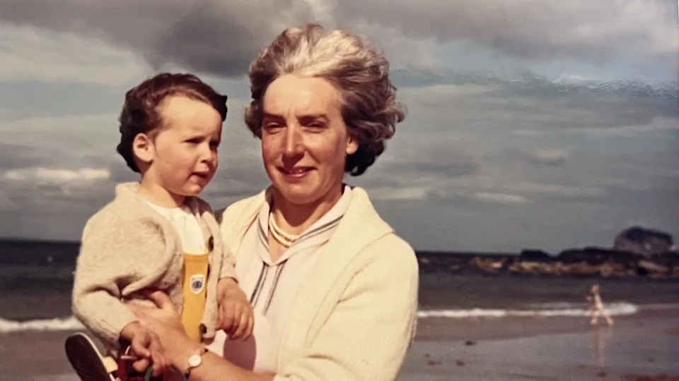Alex Morgan as a child being held by her mother on a wind-swept beach. Both are wearing a cream cardigan and their hair is blowing in the wind.