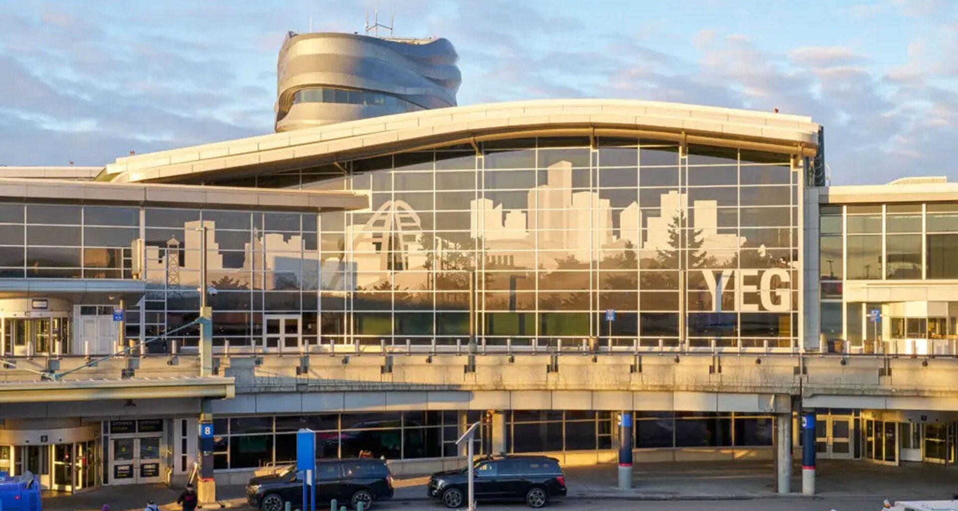 Exterior view of Edmonton International Airport departures terminal, featuring a large glass facade with a city skyline graphic and the letters “YEG” displayed at sunset.