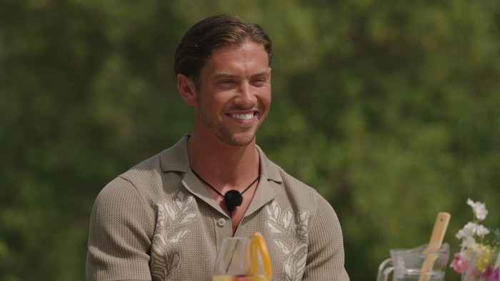 Love Island S13 A man wearing a light brown polo shirt smiling ahead looking at someone ahead of him.