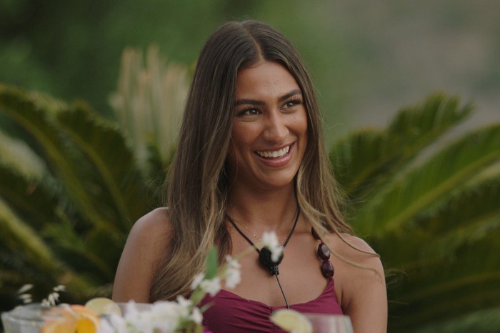 Love Island S13 Priya sat down at a table smiling ahead as she looks up at someone.