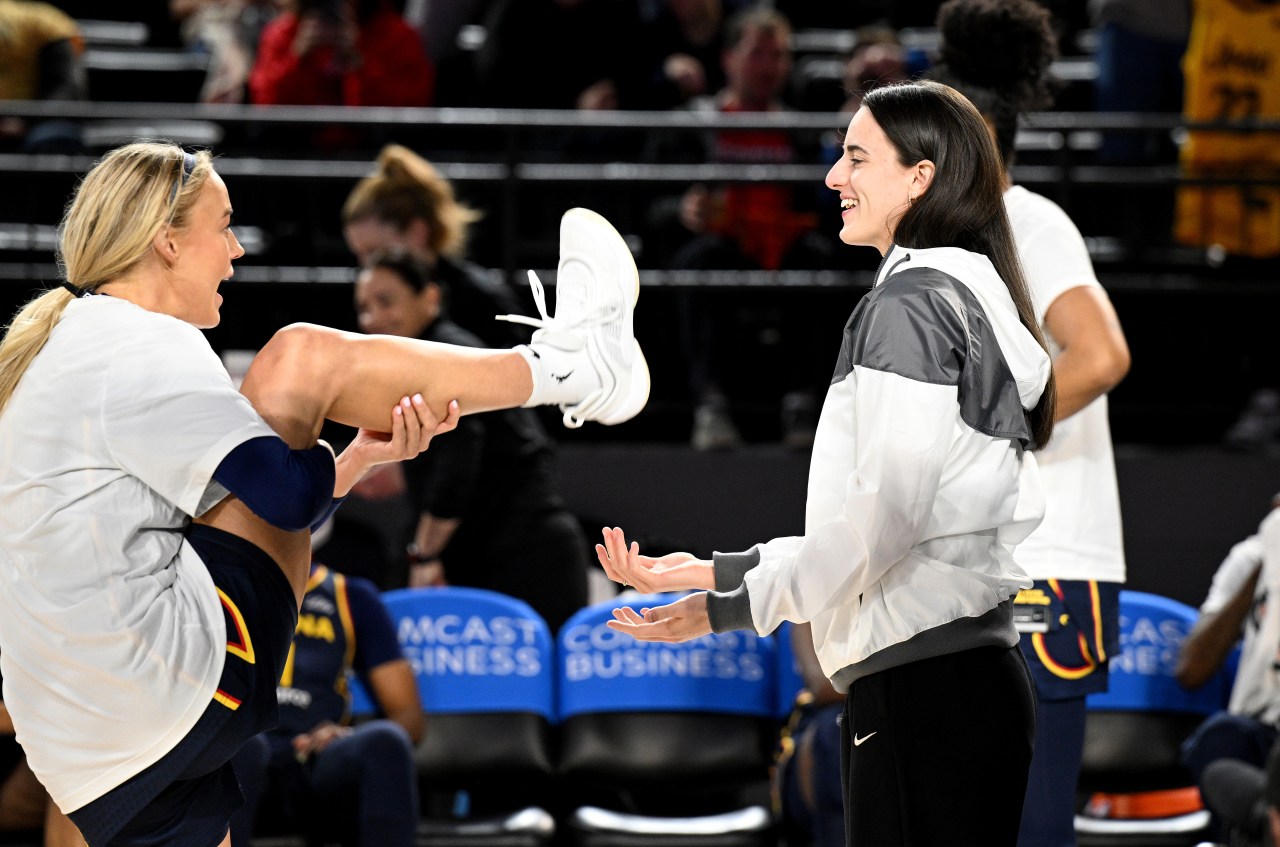 Two women in white warmup gear laugh together on a basketball court, one kicking her leg up while the other smiles, with arena seats in the background.