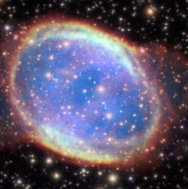 A telescope image of a planetary nebula, showing a nearly circular shell of glowing gas surrounding a blue interior filled with scattered stars. The outer ring is rendered in vivid shades of red, orange, and yellow, while the inner region glows in soft blue and teal. Background stars of various colors are visible around the nebula against a dark sky.