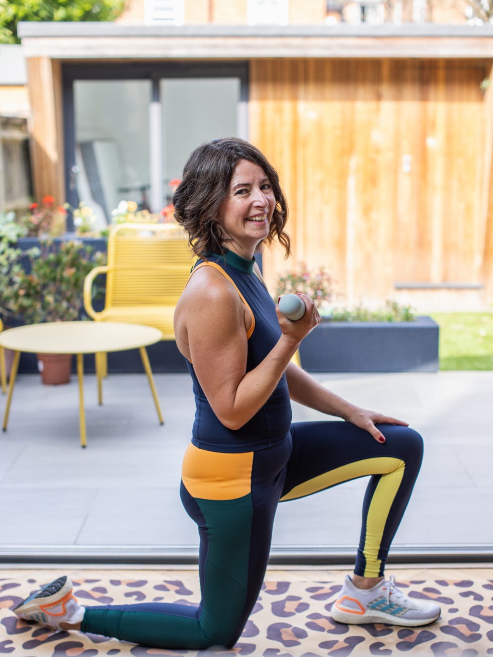 woman in workout attire kneeling with a dumbbell indoors.
