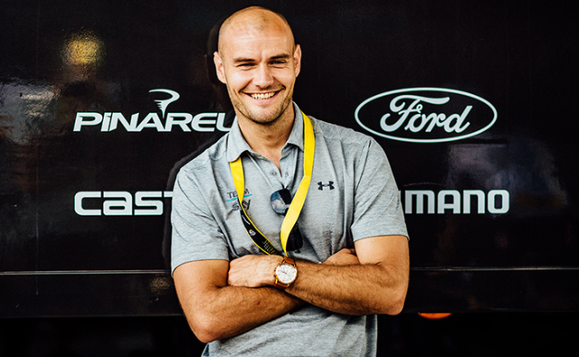 smiling man with a bald head wearing a gray polo and yellow lanyard, standing in front of a branded wall.