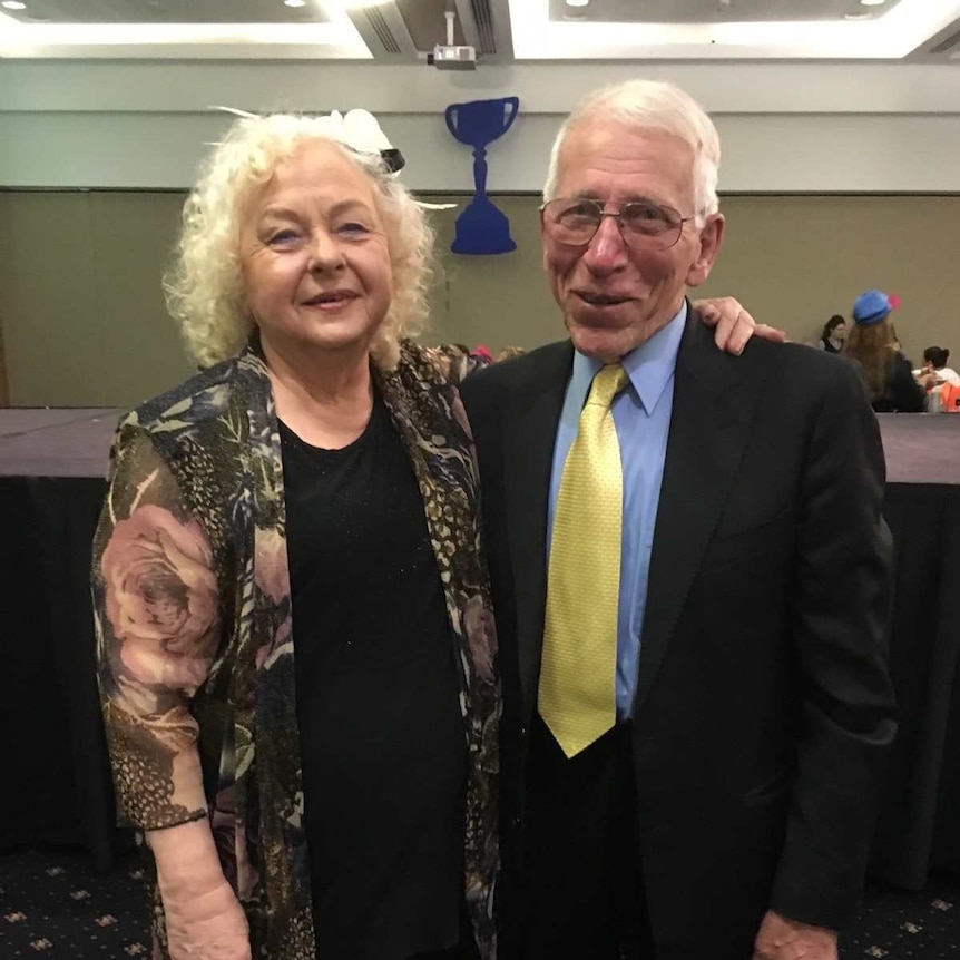 Two older people stand arm in arm smiling, wearing formal clothing.