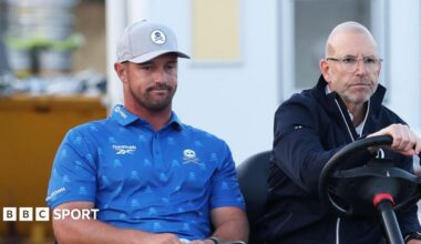 Bryson DeChambeau is driven on a buggy by an Open rules official