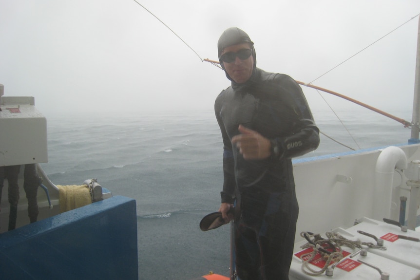 Man in full-body black wetsuit stands at side of boat. Low cloud, rain in the background.