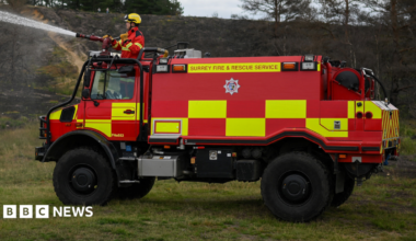 An off-road fire engine with large tires and a red and yellow livery. Surrey Fire and Rescue Service is written along the top of the vehicle. A man is spraying water from a hose above the cab.