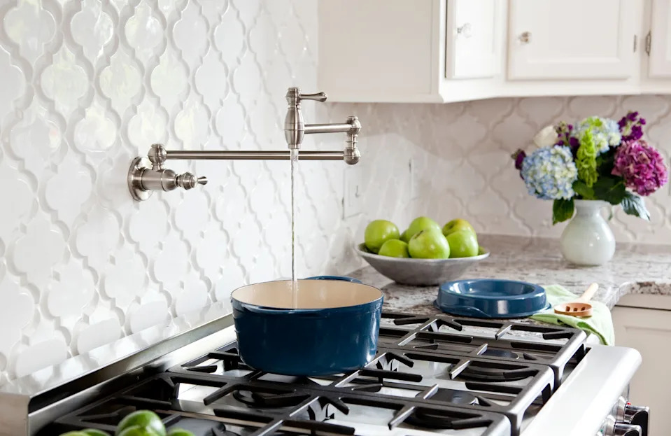 Pot filler faucet above a stove fills a blue pot. Countertop has a bowl of apples and a vase of flowers. Kitchen setting
