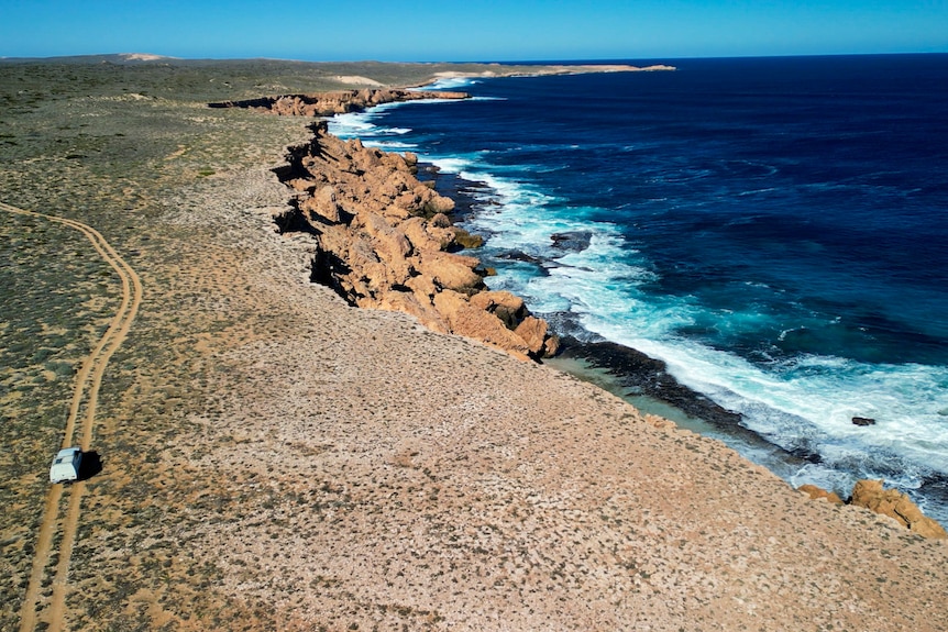 A drone image of a white ute on a dirt track, alongside a sharp island clifface.