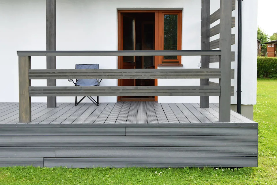 A modern, minimalistic patio with a single folding chair, overlooking a grassy yard. The porch has wooden railings and steps