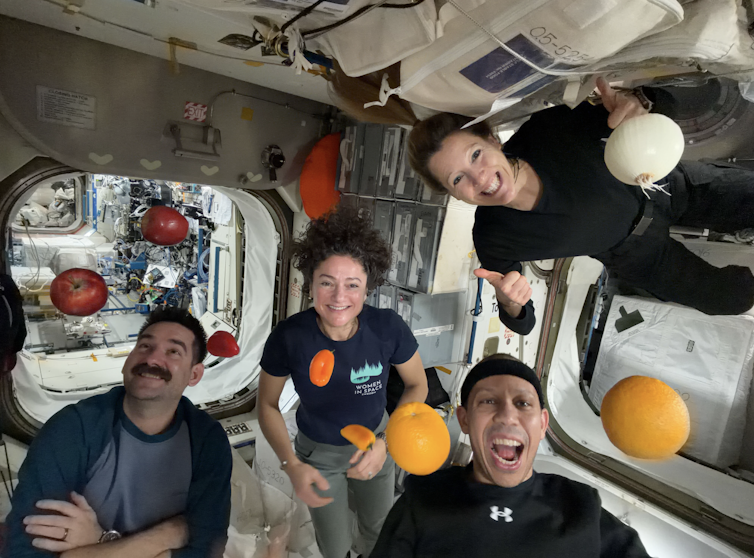 Four people floating in a small capsule, alongside several oranges and apples.