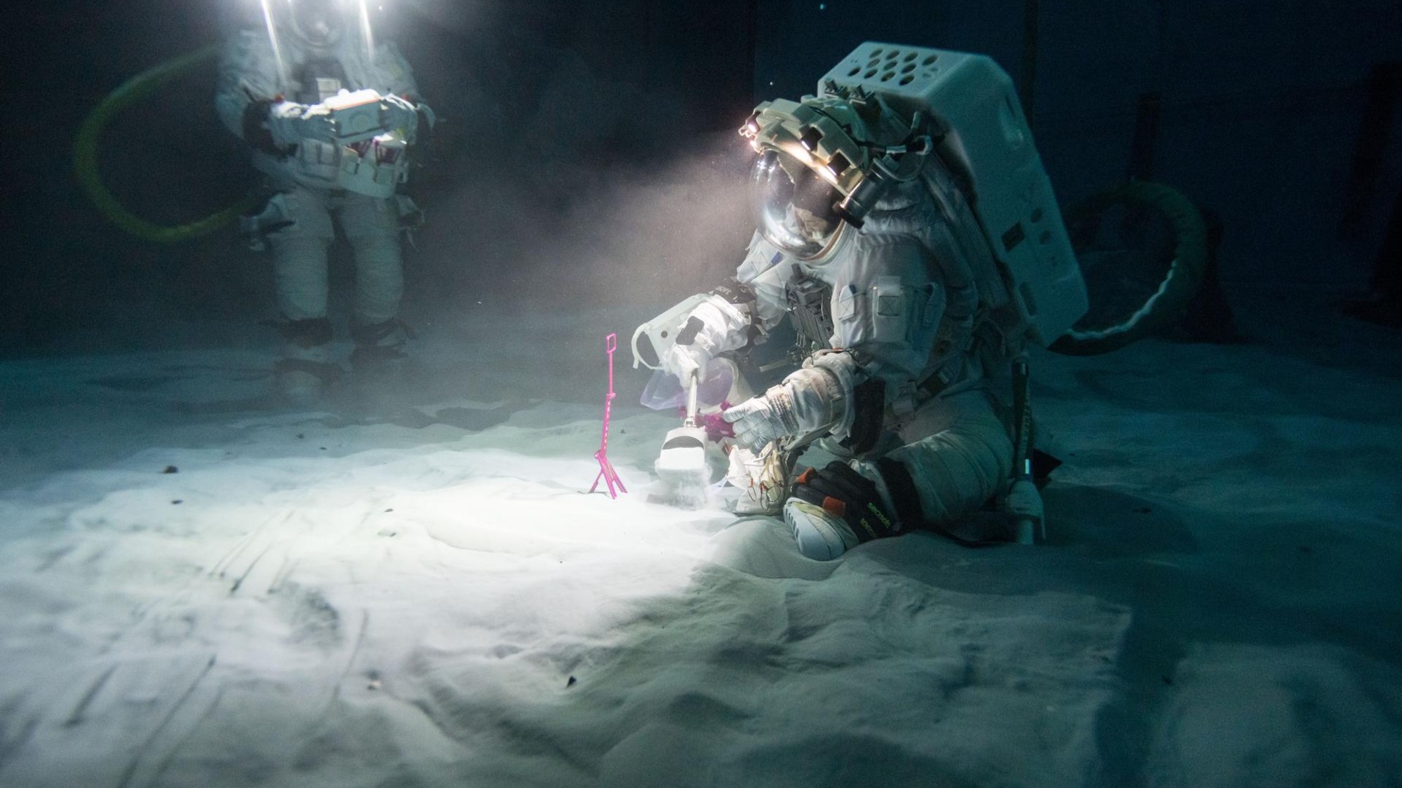 people in bulky spacesuits walk across a sandy surface underwater in a dark pool-like laboratory