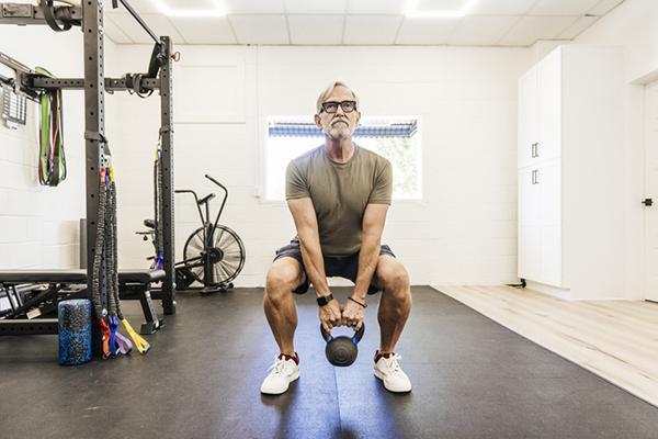 A mature man works out in a home gym, performing a squat exercise with a kettlebell.