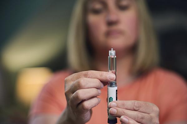 A woman holds an injector pen containing weight loss medication; the focus is on her hands and the injector, while her face is out of focus in the background.