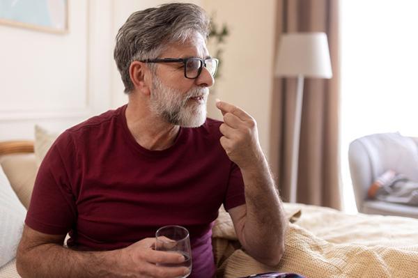 A mature man sitting on a bed takes a pill while holding a glass of water with his other hand.