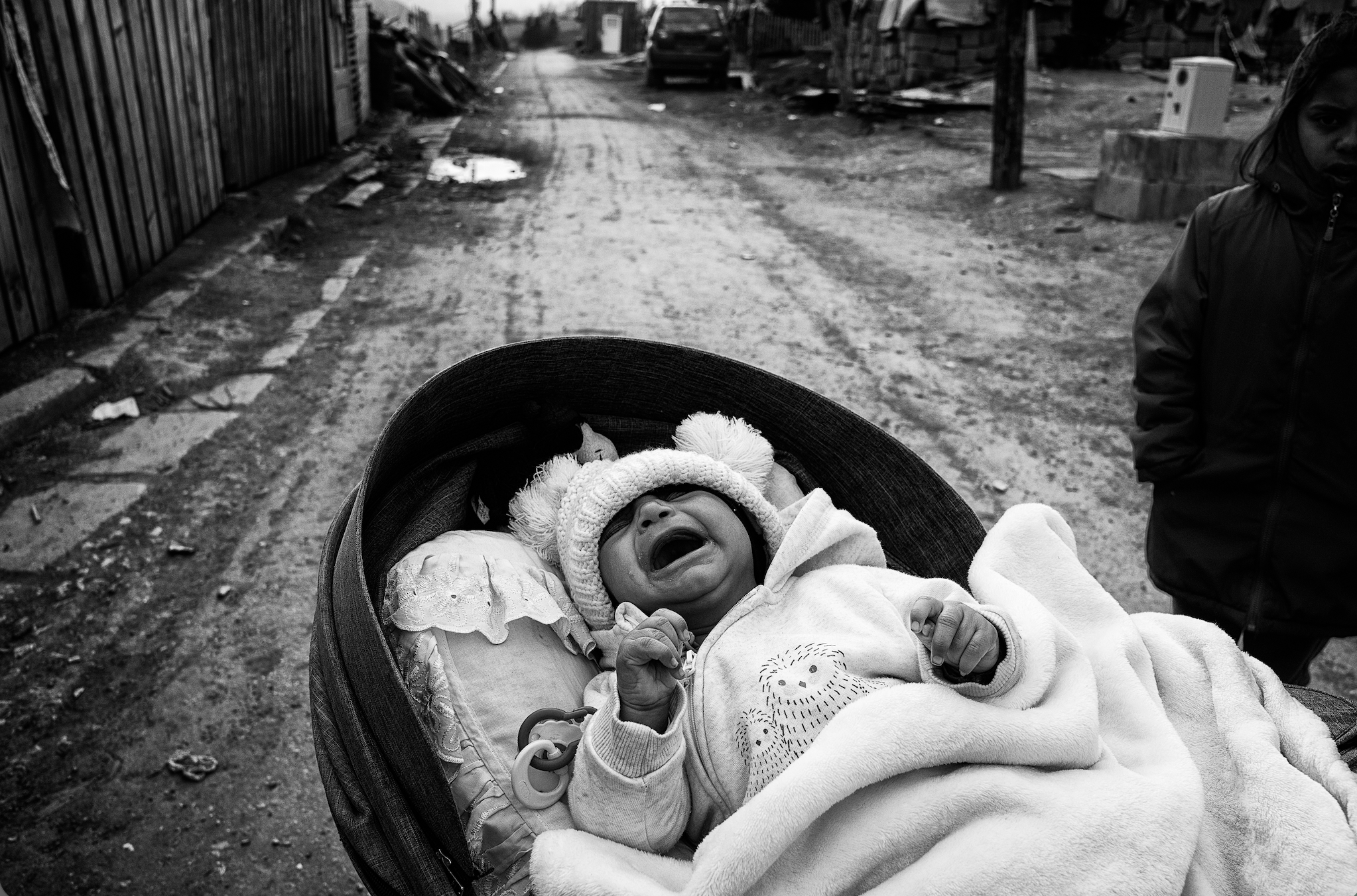 A black-and-white photograph captures a crying baby bundled in a blanket and a knit winter hat lying in a stroller, with a rural dirt road and fences stretching out in the background