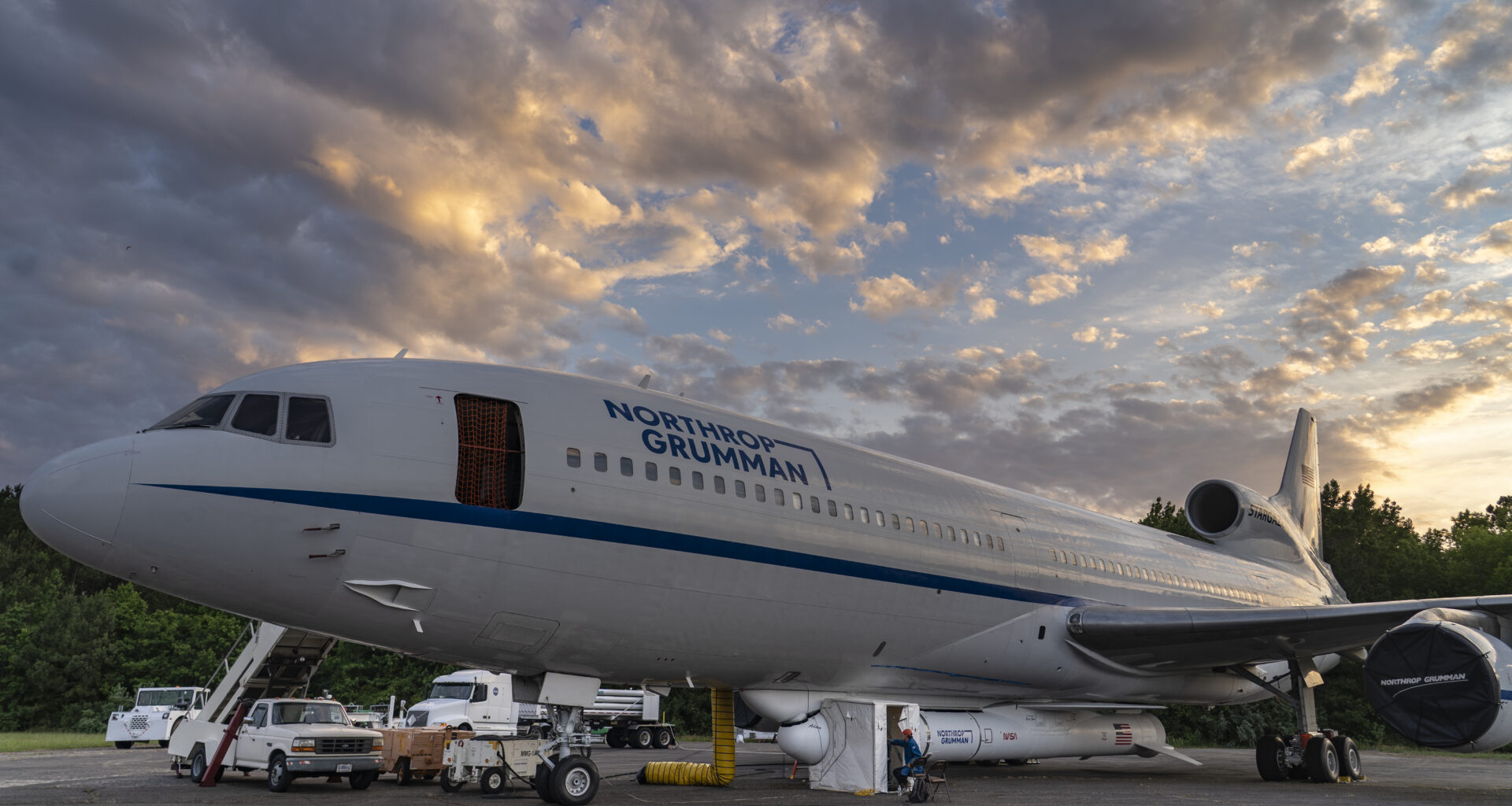 A large white airplane has a white rocket attached to the bottom of its fuselage. There is a small white popup tent covering a section of the rocket, and a person in a red ball cap and blue electrostatic discharge jacket sits in front of it. The rocket has several logos on the side and an American flag painted near the tail. The airplane has a long blue stripe running down the side, under the windows and above the wing. Blue letters spell out “Northrop Grumman” above the windows to the right of the open forward boarding door. Several support trucks and other vehicles are visible in the background. The sky is partly cloudy. 