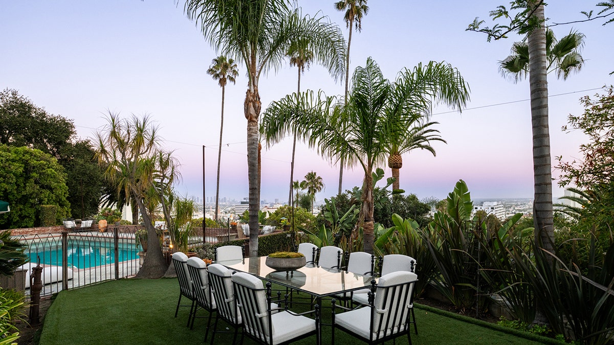 Outdoor patio seating overlooks a swimming pool with palm trees and a distant city skyline.