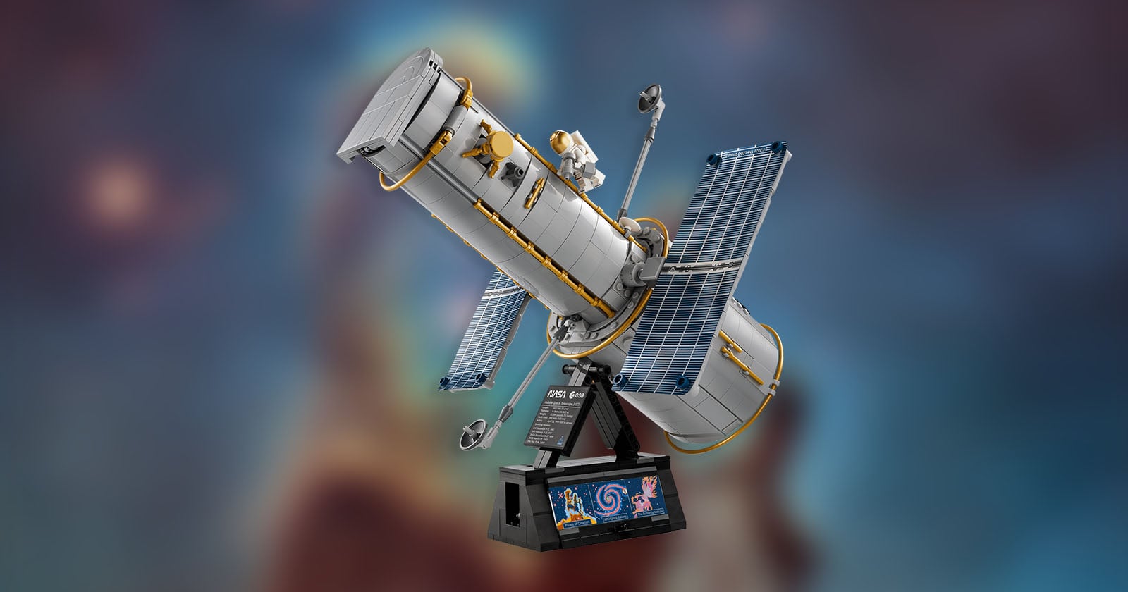 LEGO model of the Hubble Space Telescope with solar panels and mini astronaut figure, displayed on a black stand with a galaxy image, against a blurred cosmic background.