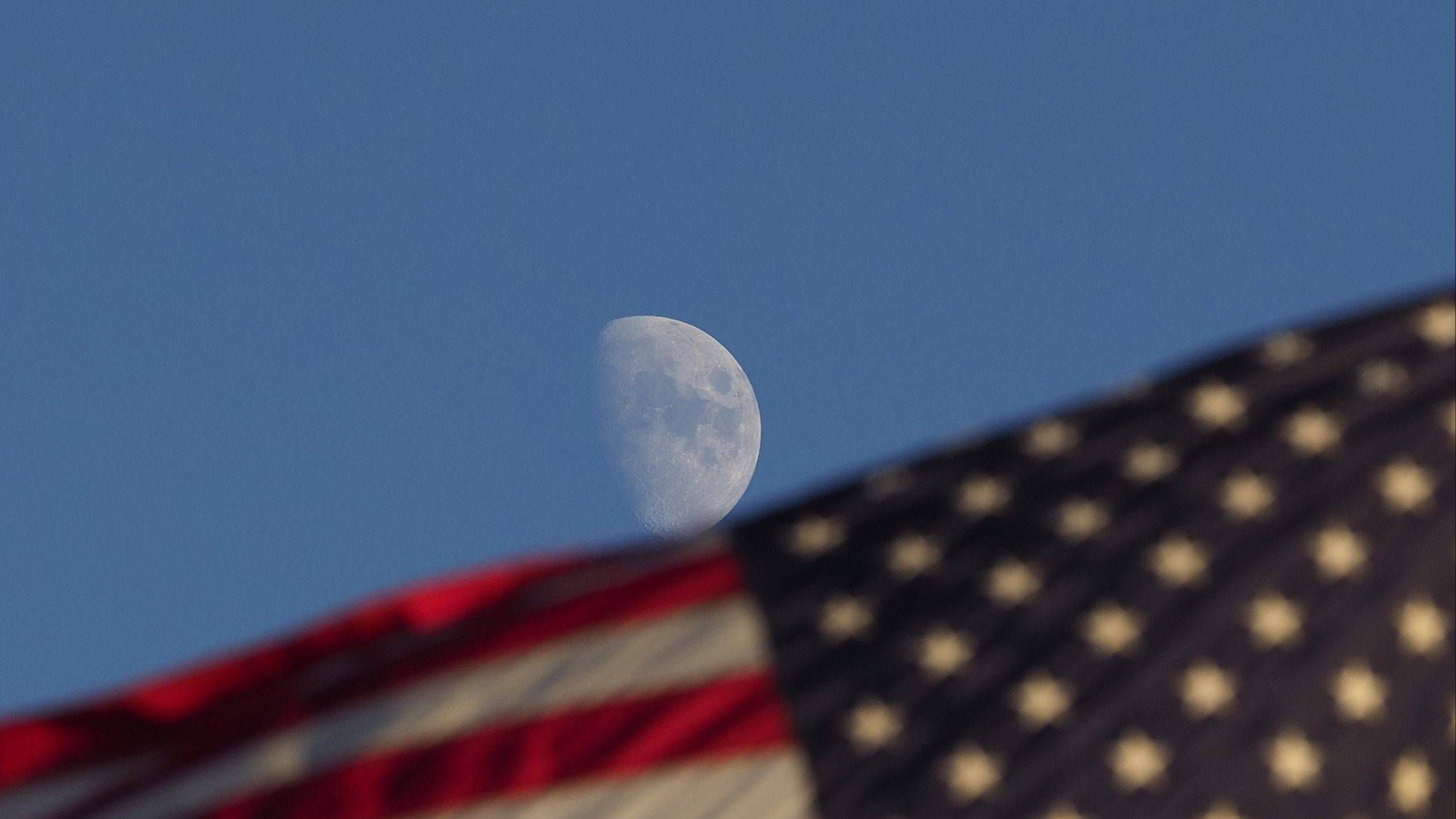 A photo showing the moon in a blue sky. On the lower half of the screen, there's an American flag.