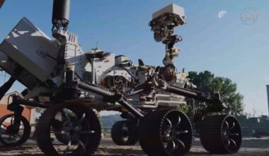 a six-wheeled rover looks up at the moon in the daytime sky.