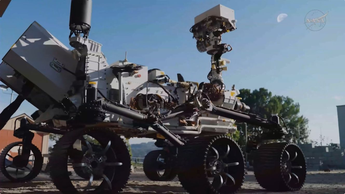 a six-wheeled rover looks up at the moon in the daytime sky.
