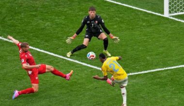 A soccer player is about to kick the ball already in the air as another player tries to stop him. The goalkeeper is ready to block.