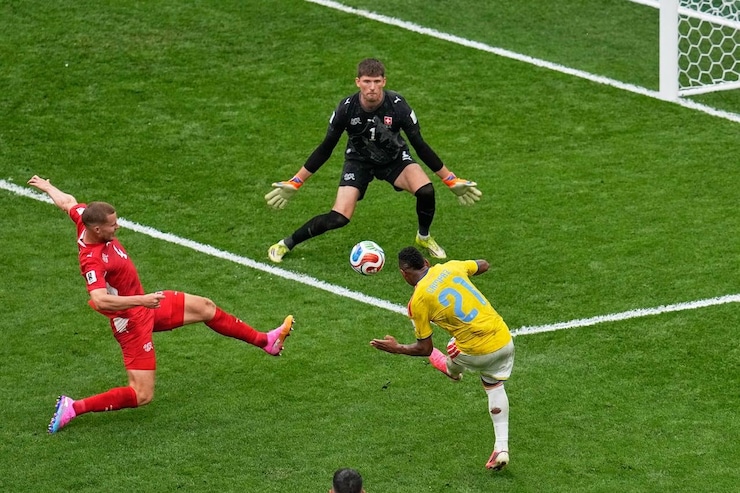 A soccer player is about to kick the ball already in the air as another player tries to stop him. The goalkeeper is ready to block.