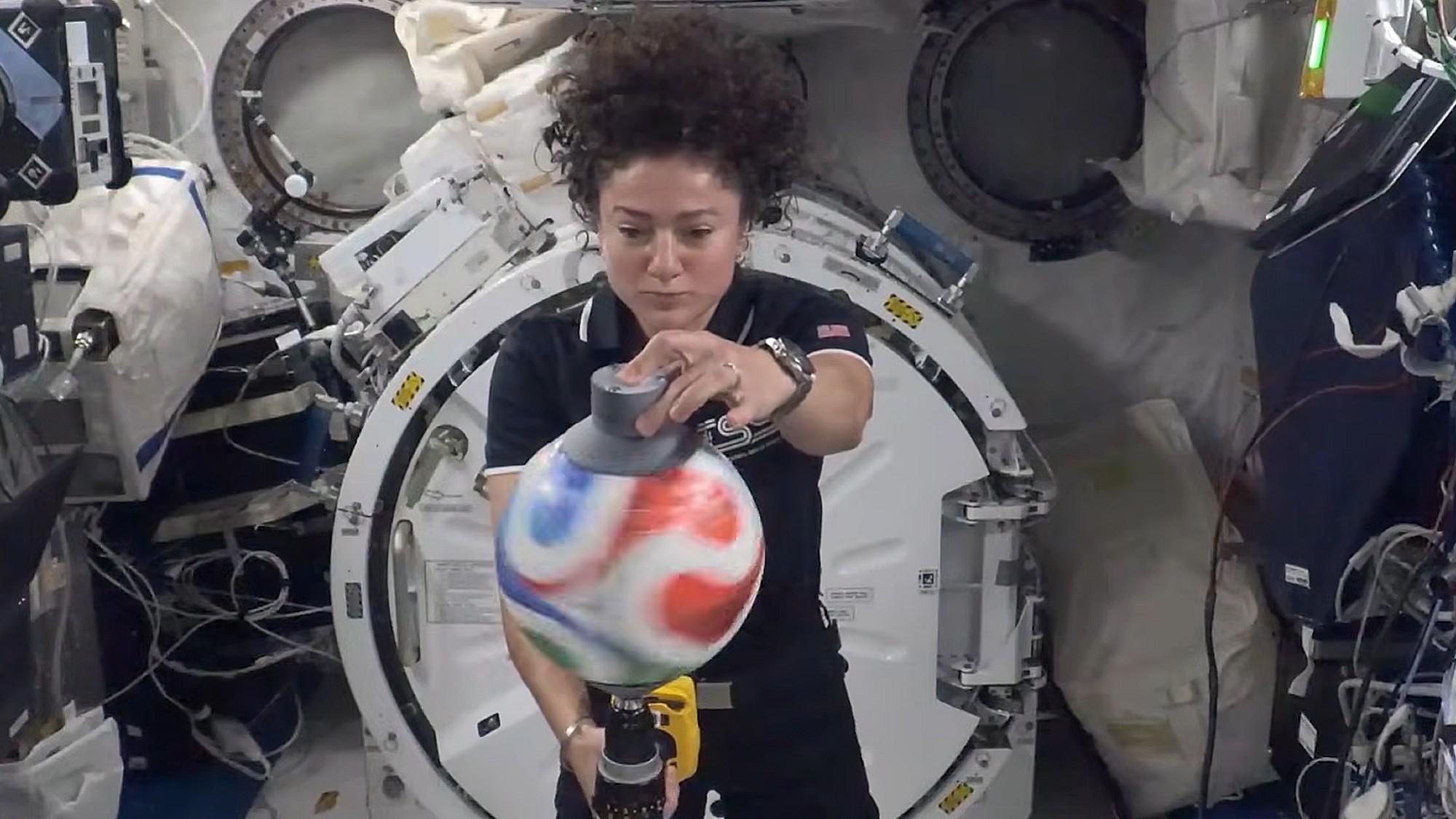 a woman floating in zero gravity in a cramped laboratory spins a soccer ball using a power drill